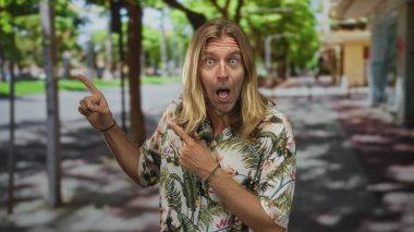 Uzun saçlı, uzun saçlı, tropikal gömlekli, güneşli, yapraklı bir kaldırım kenarında parmağını gösteren sarışın bir adam. Sürpriz..