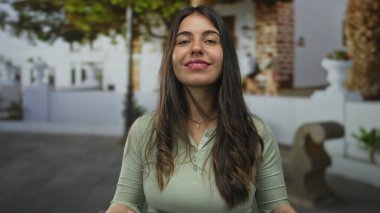 Çevresinde ağaçlar ve geleneksel mimari bulunan büyüleyici kentsel alanda gülümseyen genç bir kadın, toplum ortamında mutluluğu ve gündelik modayı somutlaştırıyor.