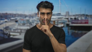 Siyah tişörtlü bronzlaşmış adam, güneşli iskele rıhtımında sessizlik için parmaklarını dudaklarına tutar; gizlilik gizlilik huzuru.