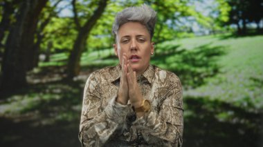 Kısa gri saçlı bir kadın yeşil ormanda soğuk ellere sürtünüyor ve üflüyor desenli gömlek giyiyor; rahatlama.