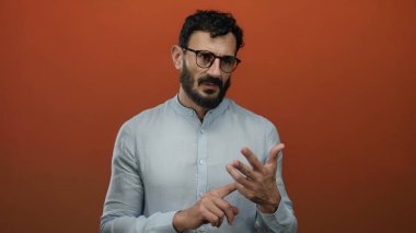 Sakallı ve gözlüklü bir adam turuncu arka planda duruyor, parmaklarına güveniyor, mavi gömlek giyiyor, İspanyol gömleği giyiyor, düşünceli bir ifade takınıyor, hareketli, soyutlanmış, yetişkin, anlamlı..