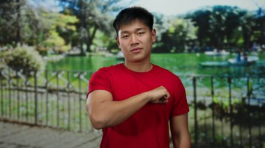 Kırmızı gömlekli genç adam yeşil ağaçlar ve dingin göl arka planıyla güneşli bir park ortamında yumruğunu uzatarak açık havada dostça bir hava yaratıyor..