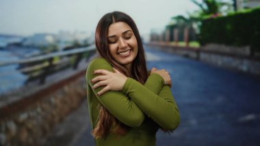 Güneşli bir sahil yürüyüşünde gülümseyen bir kadın okyanusun arka planında neşe ve dinginlik sergiliyor.