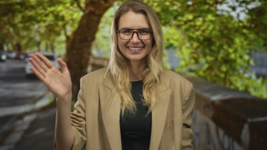 Gülümseyen ve el sallayan bir kadın gözlüklü ve bej ceketli bir kadın. Benekli güneş ışığı altında, açık havada sıcak ve rahatlatıcı bir gün olduğunu gösteriyor..