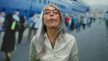 Gri saçlı yaşlı bir kadın limanda komik bir surat yapıyor, deniz kenarında bir yolcu gemisinin yanında duruyor, denizcilik ortamında neşe ve neşe ifade ediyor..