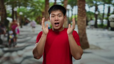 Kırmızı tişörtlü genç Çinli adam, bulanık insanlar ve gür ağaçlarla çevrili canlı bir park ortamını görünce şaşırdı, neşeli bir açık hava anını yakaladı..