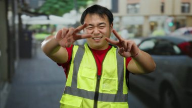 Yüzünde yansıtıcı yelek giymiş gülümseyen Asyalı bir adamın şehir caddesinde barış işareti göstermesi dostluk ve yakınlık göstergesi..