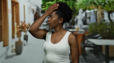 Şehir restoranındaki terasta endişeli bir kadın, dışarıda kolsuz gömlekle saksıların ve masaların arasında dikilirken düşünceli bir şekilde alnına dokunuyor..