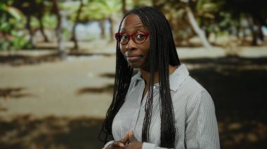 Güneşli bir parkta, uzun saçlı ve gözlüklü bir kadın. Üzerinde çizgili bir gömlek, yemyeşil ve arka planda gölgeler arasında dalgın dalgın duruyor..