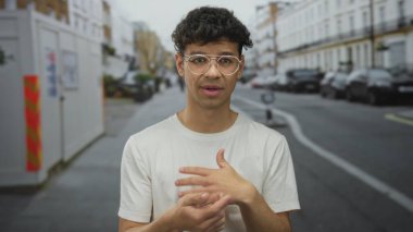 Beyaz tişörtlü, elleri çenesinde gözlüklü bir adam işlek bir şehir caddesinde duruyor, belirsizlik yansıması..