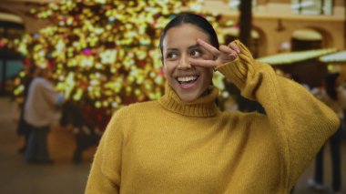 Şehir ışıklarıyla çevrili, rahat sarı bir kazak giyen açık hava kutlamalarında neşe ve tarz sergileyen bir kadın dışarıda barış jesti yapıyor..