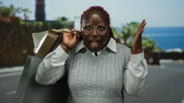 Kumsal girişine şaşıran bir kadın, güneşli bir günde elinde okyanus ve palmiye ağaçlarıyla birlikte açık havada alışveriş torbaları tutuyor..