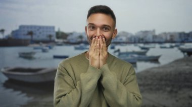 Genç İspanyol adam arka planda teknelerle deniz kenarındaki gezinti güvertesine karşı ağzını kapayarak gizli bir açık hava reaksiyonu gösteriyor..