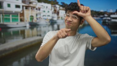Beyaz tişörtlü ve gözlüklü bir adam limanda alnına jest yapıyor. Görünür, güler yüzlü, şakacı..