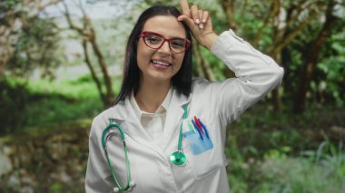 Genç İspanyol doktor, güneşli bir açık hava parkında, beyaz bir üniforma ve steteskop giyip, doğanın ortasında neşeli bir ruh hali içinde zavallı bir jest yapıyor..