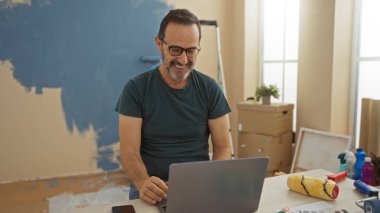 Yeni oturma odasında gülümseyen bir adam kutularla ve boya malzemeleriyle çevrili, sıcak bir evde yeni bir başlangıç öneriyor..