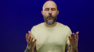 Sakallı ve dövmeli bir adam mavi bir duvara karşı meditasyon yapıyor..