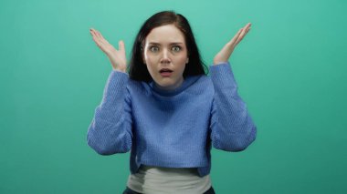 Şaşkınlık ve şaşkınlık ifade eden bir kadın canlı yeşil bir arka plana karşı, mavi bir kazak giyen, esmer oyuncu ve meraklı duyguları vurgulayan, geniş gözlü, eller kaldırılmış.