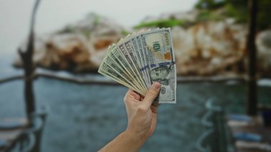 Manzaralı bir sahil sahilinde Amerikan doları tutan adam, açık havada güzel bir sahile karşı banknotlar sergiliyor..