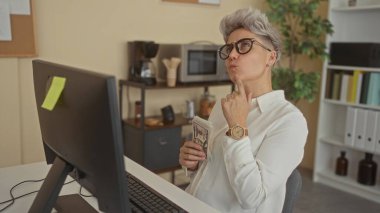 Modern ofis içlerinde bilgisayar monitörüyle nakit para destesi tutan gözlüklü kısa saçlı kadın; bütçe finansmanı stresi.