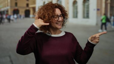 Elle işaret veren bir kadın, canlı bir şehir caddesinde duruyor, gülümsüyor ve gözlük takıyor..