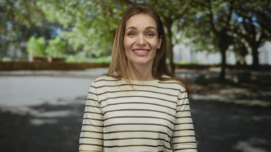 Parlak güneş ışığı altında yeşil parkta kolları açık gülümseyen çizgili tişört giyen genç bir kadın..