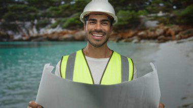 Beyaz kasketli ve yansıtıcı yelekli bir adam ellerinde taslak okuyor ve inşaat sahasında plajdan sahile çalışma planı yapıyor. İyimser planlama..