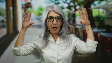 Canlı bir sokak sahnesinde bir açık hava restoranının terasında enerjik bir şekilde konuşurken gri saçlı yaşlı bir kadın el hareketi yapıyor..