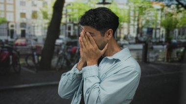 Akşam karanlığında kanal kenarında kalabalık bir şehir sokağında kot ceketli bir adam yüzünü kapatıyor..