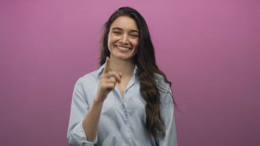 Mavi çizgili tişörtlü genç İspanyol kadın gülümsüyor ve pembe stüdyoyu işaret ediyor..
