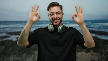 Sakallı ve kulaklıklı genç adam deniz kenarında neşeli bir ifadeyle rahatlamış ve kaygısız bir havayı somutlaştırıyor..