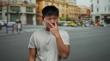 Şehir caddesinde ihtiyatlı bir jest yapan genç adam, arka planda binalar ve bulanık yayalarla canlı şehir yaşamını ve kültürel çeşitliliği vurguluyor..
