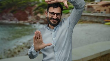 Sahilde sakallı ve gözlüklü bir adam ellerini deniz manzarasını çerçeveletip doğada yaratıcılık ve neşe ifade ediyor..