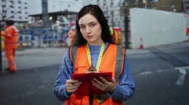 Esmer saçlı bir kadın, inşaat alanında gönüllü olarak turuncu bir yelek giyiyor, elinde bir pano var, etrafı parlak giyinmiş işçiler tarafından çevrilmiş, sitenin düzenbazlığını yakalıyor..