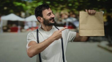 Arka planda bulanık market tezgahları ve yeşil ağaçlarla çevrili, gösterirken memnun görünen, bir sokakta kağıt bir çanta tutan İspanyol adam..