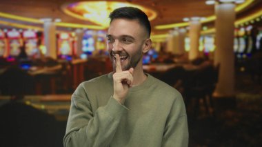 Genç adam kumarhane ortamında canlı oyunlar ve renkli ışıklarla çevrili, gülüyor ve yüzünde neşeli bir ifadeyle şşşş hareketi yapıyor..