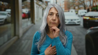 Gri saçlı, mavi kazaklı yaşlı bir kadın arka planda arabalarla dolu bir şehir sokağında dalgın dalgın duruyor..