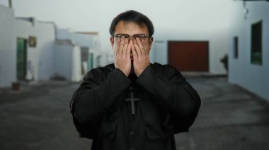 Gözlüklü ve haçlı siyah cüppeli Asyalı bir adam şehir caddesinde duruyor, sakin bir kasaba ortamında düşünce ve yansıma hissi yayıyor..