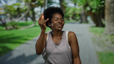 Gri elbiseli kadın, güneşli park ortamında taştan bir jest yapıyor, kendine güveni tam, yemyeşil ve ağaçlarla çevrili, genç açık hava enerjisini somutlaştırıyor..