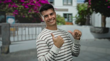 Gülümseyen ve el kol hareketi yapan genç bir adam canlı bir şehir caddesinde çiçekli bir arka planda gün ortasında çekilen çekimlerde neşe ve pozitiflik saçan.