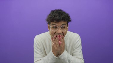 İspanyol adam mor stüdyo ortamında elleri ile ağız kaplar kameraya bakarken gözlük ve beyaz gömlek giyer; sessizlik.