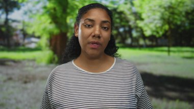 Çizgili gömlekli kadın başını eğiyor ve parktaki yeşil ağaçların arasında yüzünü gösteriyor..