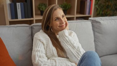 Oturma odasında rahat bir kadın rahat bir süveter giyiyor ve kanepede düşünceli bir yüz ifadesi sergiliyor. Gün içinde rahat bir hava sergiliyor..