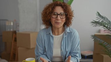 Oturma odasında gülümseyen bir kadın. Etrafı kutularla çevrili. Yeni ev heyecanını yansıtıyor..