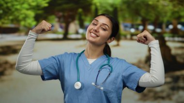 Önlüklü İspanyol kadın güneşli bir parkta esnek kollarıyla güven içinde poz veriyor. Açık hava sağlık hizmetlerinde kuvvet ve pozitifliği somutlaştırıyor..