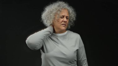 Gri saçlı bir kadın koyu bir arka plan karşısında boyun ağrısı çekiyor, izolasyonda rahatsızlık ve stres taşıyor..