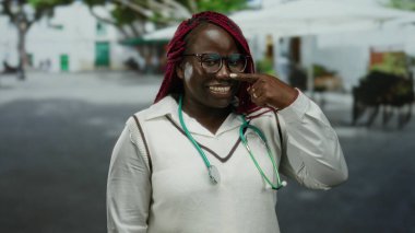 Sokakta steteskoplu gülümseyen kadın gözlüklü ve örgülü saçlarıyla burnuna bakıyor. Arkadaşça ve cana yakın bir tavır sergiliyor..