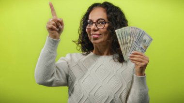 Gözlüklü orta yaşlı bir kadın, Amerikan doları banknotlarını canlı sarı bir arka plana karşı tutuyor, zenginlik ve güveni resmediyor..