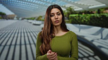 Yeşil tişörtlü, uzun saçlı genç bir kadın açık havada modern mimari geçmişi ve doğal ışıklandırmasıyla sakin bir şehir parkı ortamında duruyor..