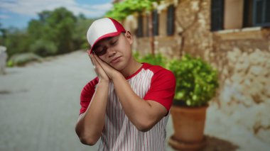 Beyzbol üniformalı genç bir adam şehir caddesinde bir uyku jesti yapıyor, canlı bir şehir ortamında yorgunluk gösteriyor..